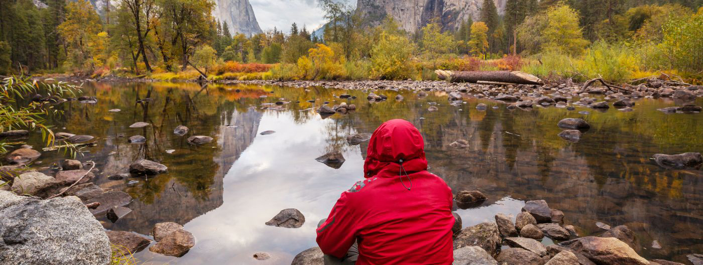 Person in a red jacket and hood sitting in Yosemite National Park with their back to the camera