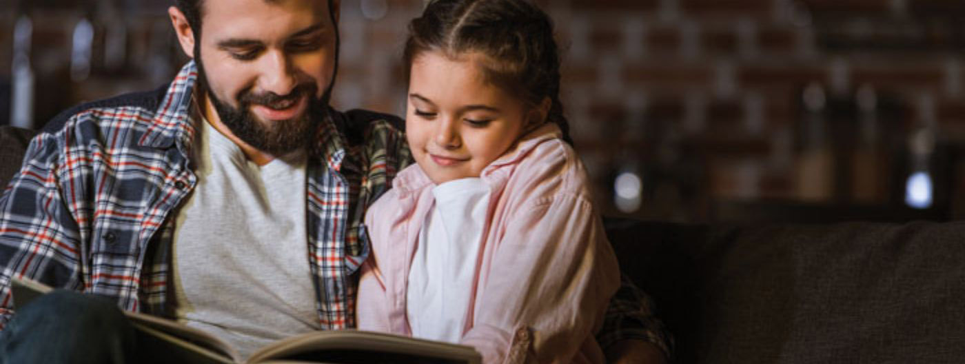 Bearded father in flannel shirt reading a book to his daughter next to him on a couch.