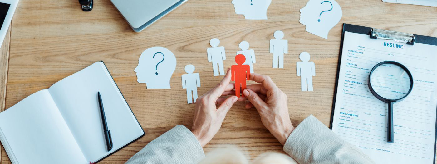 Table viewed from above with a woman holding a red paper cutout of a figure, surrounded by white cut-out figures, a cut-out head with a question mark, a form with a magnifying glass, and an open notebook.