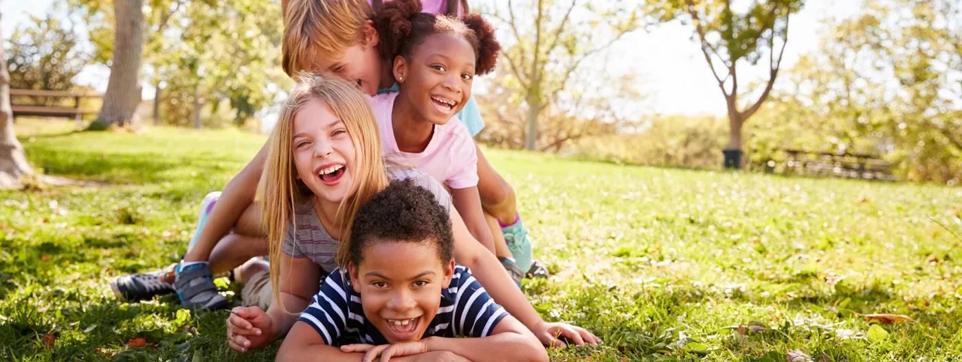 A group of smiling children form a pyramid by lying on top of each other in a sunny, grassy setting