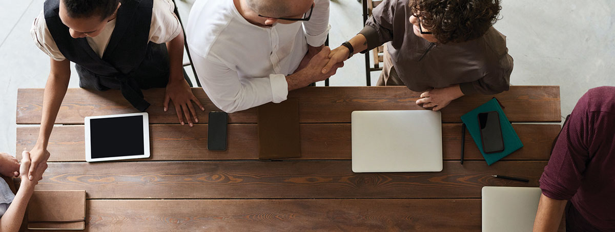 Men and womens seen from above, standing around a table with laptops and tablets, shaking hands